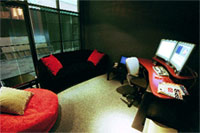 A dark room featuring a black sofa with red pillows, a red desk with dual monitors, and a red circular ottoman.