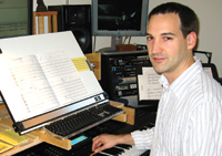 A man in a striped shirt sits at a desk with sheet music, a keyboard, and computer monitors.