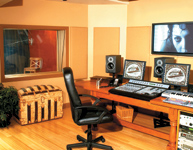 A recording studio control room with a large mixing console, monitors, computer screens, and a view into a booth.