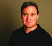 A man with short dark hair wearing a black polo shirt against a plain dark background.