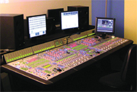 A large audio mixing desk with numerous sliders and buttons, surrounded by computer monitors and speakers.