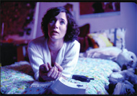 A woman with dark curly hair lies on a patterned bed, looking up with a worried expression.