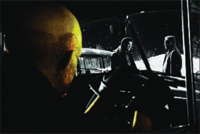 A close-up profile of a person looking out a window at two figures standing by a car in the dark.