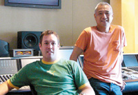 A man in a green shirt sits while a man in an orange shirt stands in a recording studio.