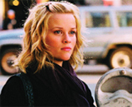 A blonde woman with wavy hair wearing a dark scarf looks into the distance on a city street.