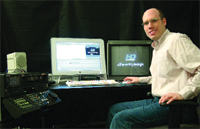 A man in a light shirt sits at a desk with two monitors and audio mixing equipment.