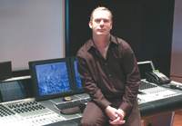 A man in a dark shirt sits behind a large audio mixing desk with multiple monitors in a studio.