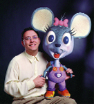 A man in a light yellow shirt sits next to a large, cartoonish grey mouse puppet with pink ears and a bow.