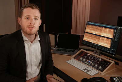 A man in a black blazer and white shirt sits at a desk with a laptop, monitor, and audio mixing console.