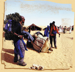 Two travelers stand near a resting camel in a sandy desert landscape with tents in the background.