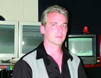 A man with light hair wearing a black shirt and grey vest, standing in front of several computer monitors.