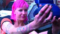 A woman with vibrant pink hair and tattoos on her arm holds a steering wheel while driving.