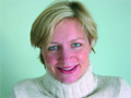 A blonde woman smiling while wearing a white turtleneck sweater against a light green background.