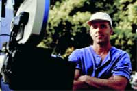 A man in a blue shirt and white cap stands with arms crossed next to a large piece of machinery.