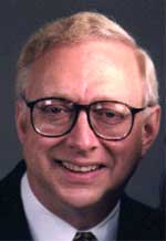A smiling man with short blond hair and black-rimmed glasses wearing a dark suit and white shirt.