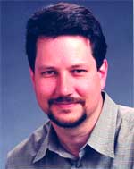 A man with dark hair and a goatee wearing a grey collared shirt against a blue background.