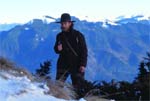 A man wearing a dark jacket and a wide-brimmed hat stands before blue mountains.