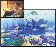 A man sits at a desk next to a large illustration of a coral reef with a sea turtle and fish.
