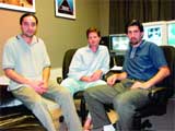 Three men sitting in chairs in a room with computer monitors in the background.