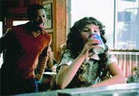 A young girl with dark curly hair drinks from a blue and white cup while a man looks on in the background.