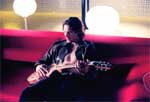A person sits on a red sofa playing an electric guitar under bright, glowing lights.