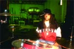 A person in a black cap and red shirt sits behind a drum kit in a room with green lighting.