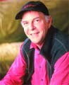 A smiling man wearing a black baseball cap, a pink button-down shirt, and a dark vest.