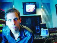 A man with short hair looks forward in a room filled with monitors and audio equipment.