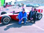 Two men squatting next to a red open-wheel racing car in a parking lot.