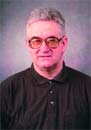 A man with gray hair and glasses wearing a black polo shirt against a purple background.