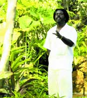 A man with dark hair wearing a white shirt and pants stands amidst dense green tropical foliage.