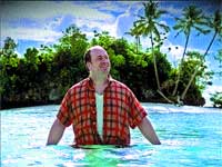 A man in a red plaid shirt smiling while waist-deep in a turquoise tropical pool with palm trees.