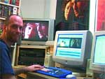 A man sits at a desk with several computer monitors displaying various images and interfaces.