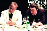 Two men in suits sit at a table with glasses and a green bottle.