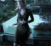 A woman in a dark dress leans against the hood of a light-colored car outdoors at night.