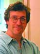 A man with wavy brown hair and glasses wearing a light blue button-down shirt.