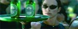 A woman wearing dark sunglasses holds a green tray with two glowing green bottles in a crowded setting.