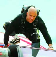 A bald man in a black long-sleeved shirt and tactical gear leans over the edge of a red vehicle.