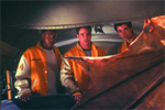 Three men in orange and white jackets look at a large, textured orange object in a dimly lit setting.