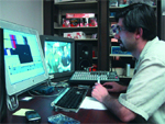 A man with glasses sits at a desk using a computer, keyboard, and mouse with two monitors visible.