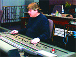 A man in a dark sweater sits at a large professional audio mixing console in a studio.