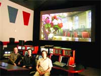 Three people sit in a dark room facing a large projection screen showing a bright interior scene.