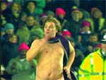 A shirtless man wearing a purple scarf stands in front of a crowd of spectators.