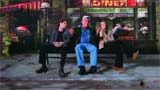 Three people sit on a bench outside a diner with neon signs at night.