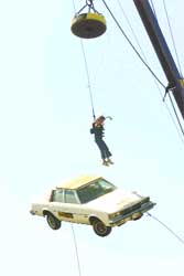 A person hangs from a cable suspended above a white car against a pale sky.