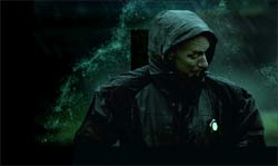 A person wearing a dark hooded jacket stands in a dark, misty setting with green light effects.