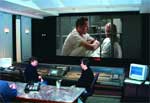 Two people sit in a dark control room facing a large screen showing two men talking.