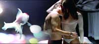 A shirtless man and a woman in a white top embrace next to a large, translucent goldfish.