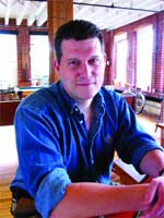 A man with short dark hair wearing a blue button-down shirt sits at a table in a room with large windows.