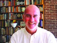 A bald man with a graying beard smiling, wearing a white button-down shirt in front of a large bookshelf.
